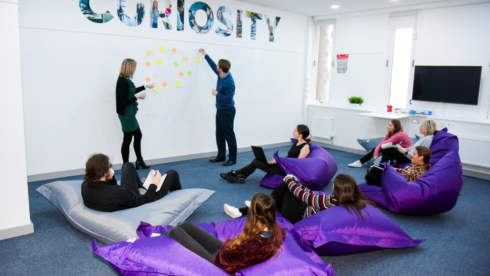 A group of people are sat on bean bag chairs on the floor of a room, while two other people put post-it notes on the wall for them to comment on