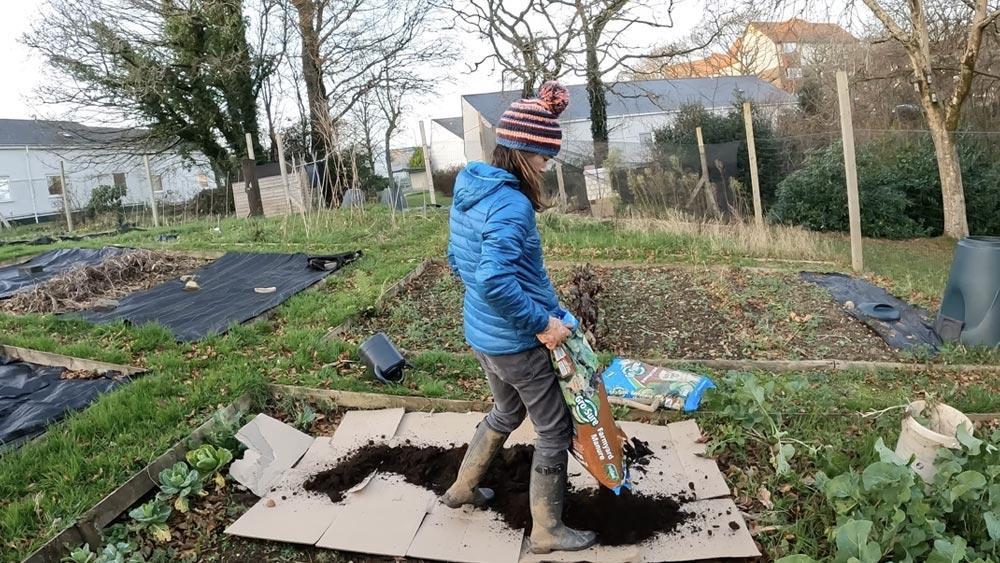 An image of a woman working on an allotment outside on the campus of Plymouth Marjon University. She is spreading a bag of farmyard manure into a vegetable bed, tending to plants. She has muddy wellington boots on, a blue puffer jacket, and a striped woollen hat with a pom pom on top.