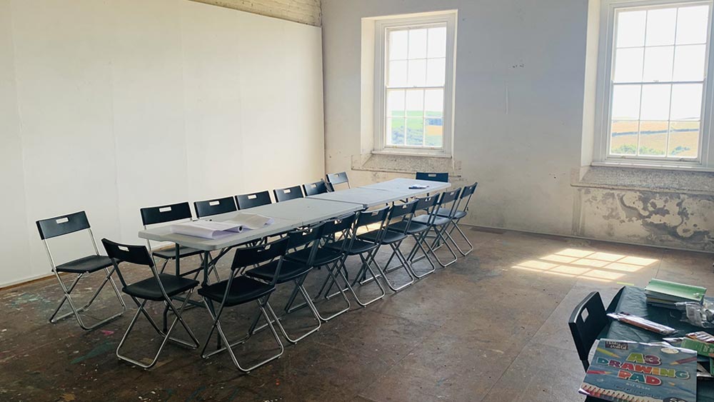 An image of a long table and chairs set neatly in an artist studio within an old military barrack building. It is a sunny day, with lots of light coming through two big windows. There is some rolled up white writing paper on the table.
