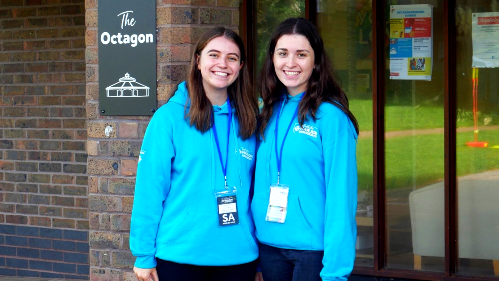 Two female student ambassadors in blue hoodies stood in front of a red brick building and smiling at the camera