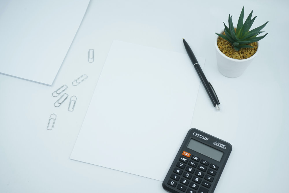 A calculator next to a piece of blank paper and a pen on a white tabletop with a cactus in a pot to the top right.
