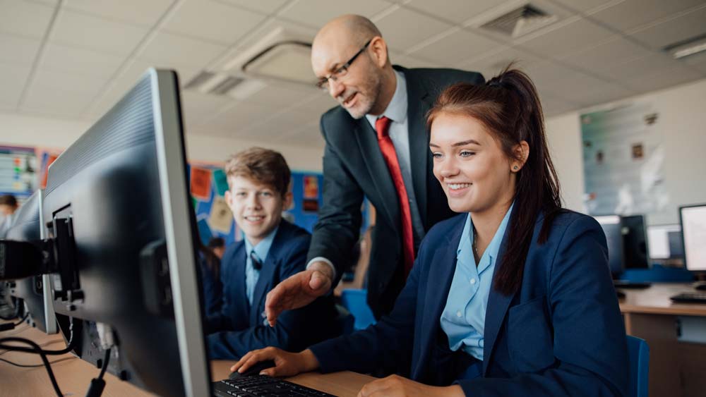 A teacher teaches two students who are doing computer work
