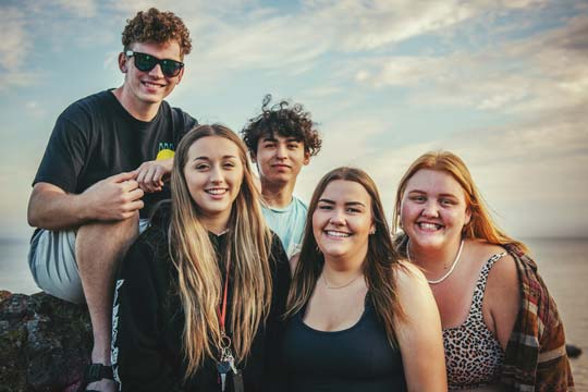 A group of five relaxed looking teenagers pose for a photo with a mellow evening sky in the background