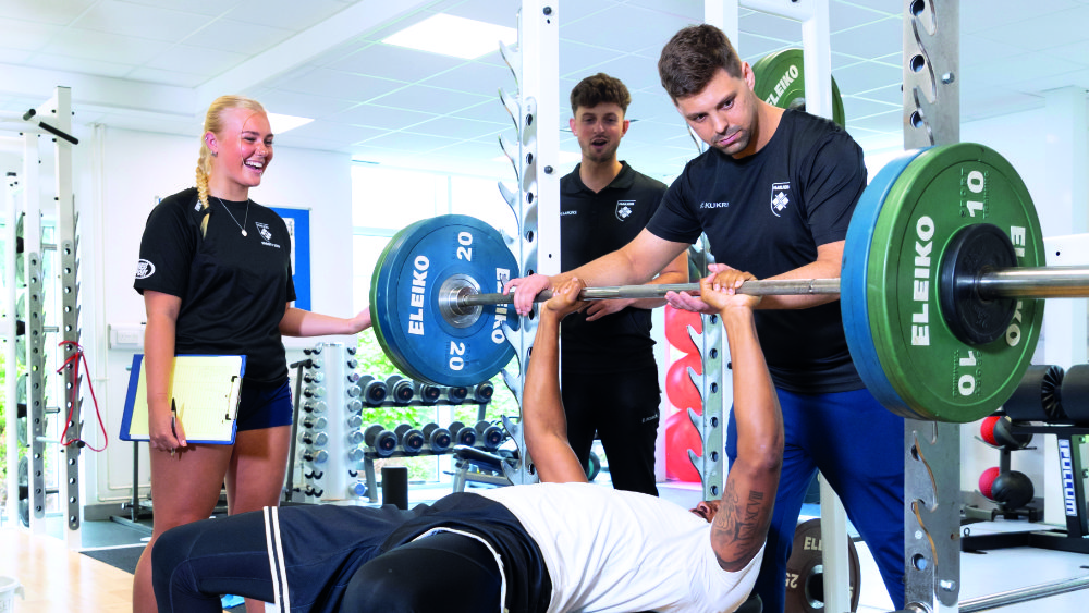 A group of Marjon sports students supervising someone benching weights in the Marjon Sport and Leisure Centre