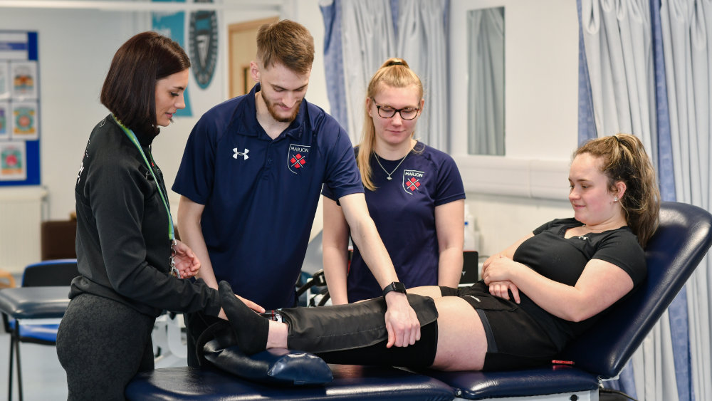 Two students are learning about injury rehabilitation by examining a patient wearing a knee brace who is lying on stretcher