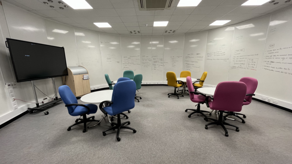 A circular room with floor to ceiling whiteboards around the wall. Four circular tables are surround by office chairs in blue, pink, yellow or green