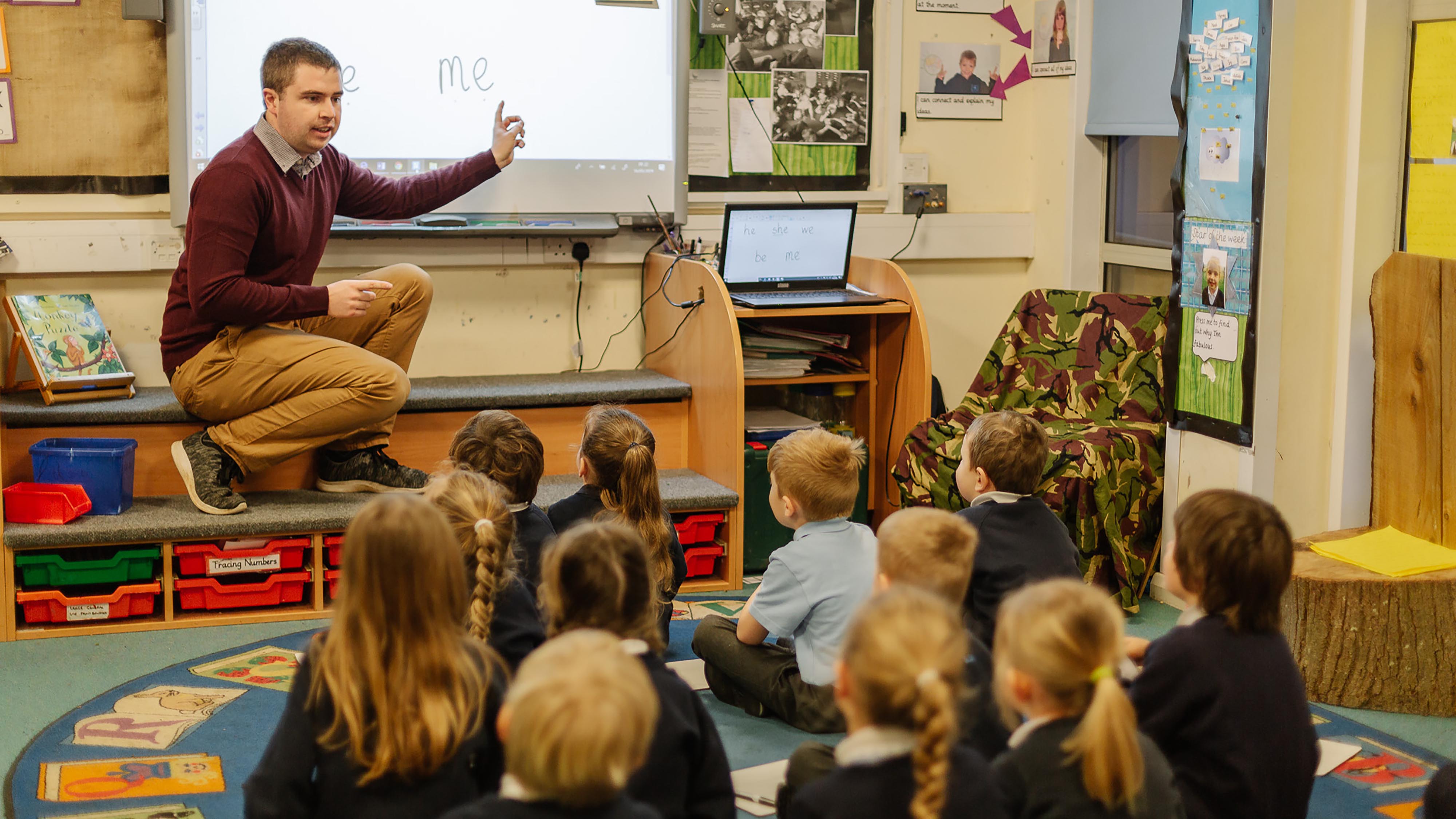 A primary school teacher talks at the board as the class look at it