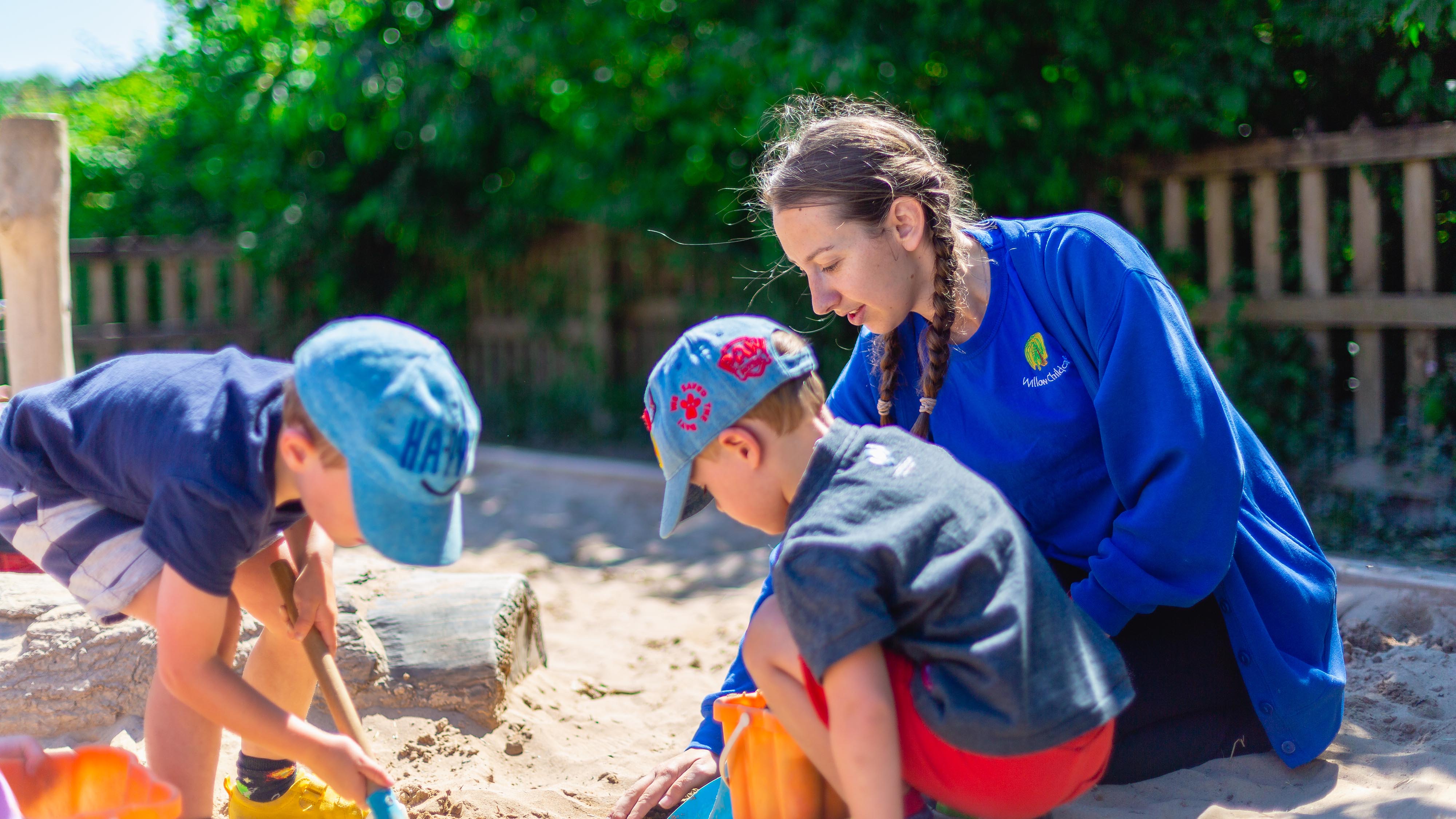 An Early Years Teacher and two toddler boys play in the sand