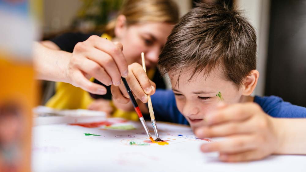 A boy with a disability paints with support from his teacher