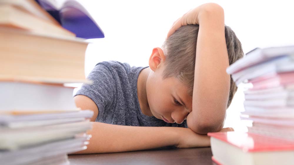 A boy is slumped on a desk, surrounded by books and looking sad