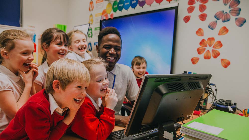A primary school teacher and his pupils are laughing whilst looking at something on the computer