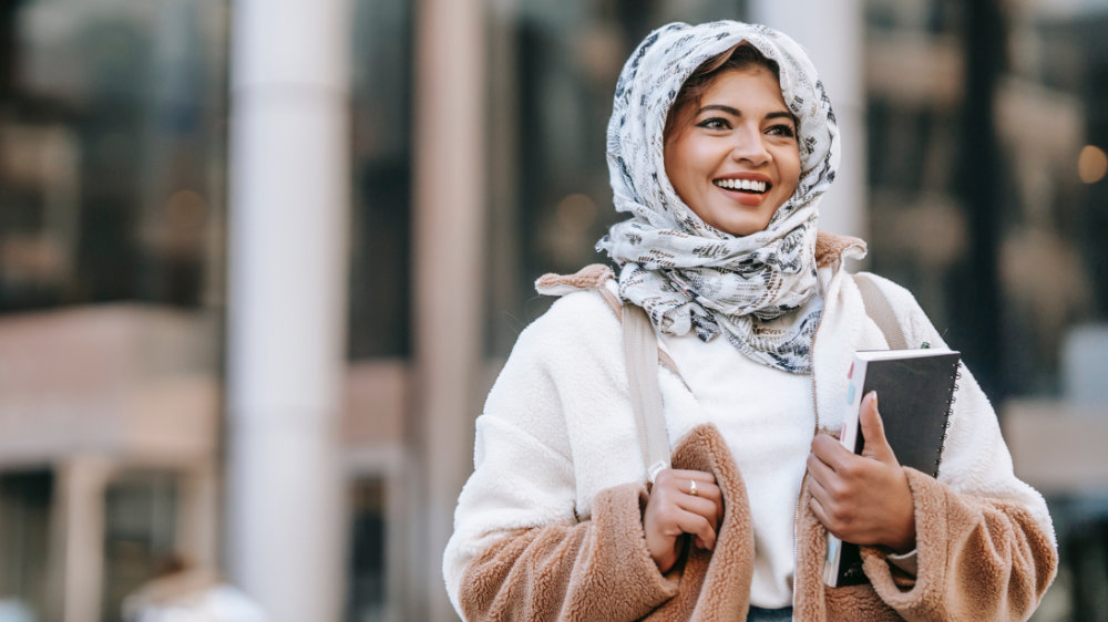 A young woman in a headscarf and chunky fleece smiles to the right of the camera