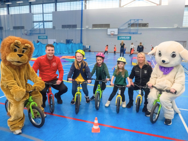 The Marjon mascots, primary school children, and Marjon staff sit on bikes in a sports hall