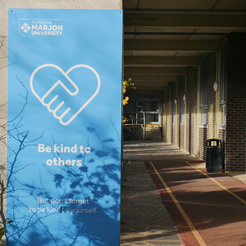 A blue sign in a Marjon walkway that reads 'Be kind to others but don't forget to be kind to yourself.'