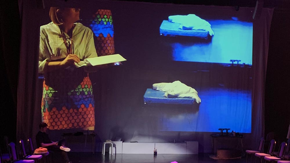 A woman in a colourful dress is holding a script in a large theatre space. Her image is projected on a large screen at the back of the room, and stage lights offer a dark and immersive atmosphere