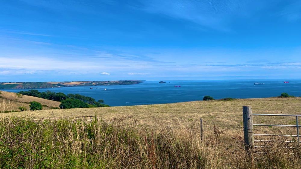 An image of the outdoor seascape across Maker Heights in Cornwall, UK taken from the top of a hill. There are fields of grasses and plants, trees, and a view of a bright blue sea. The sky is bright and there are mackerel cloud formations in the sky. Its a bright and sunny day.