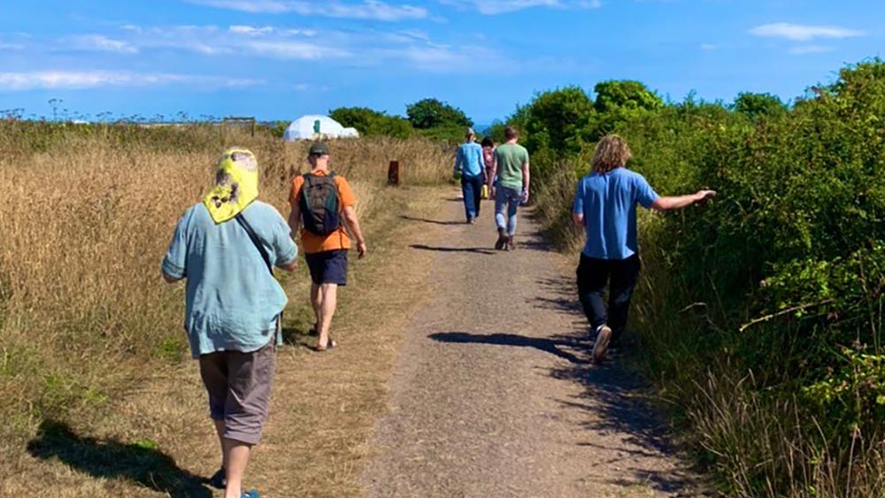 An image of a group of people, different age, sex and genders, walking down a rural path in the outdoors. There are hedgerows and tall grasses, the sun is shining and the sky is bright blue.