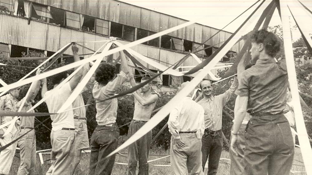 An image of a group of people in the outdoors, mixed ages, sex, and gender, holding lengths of white materials above their heads in a concentric pattern. The people in the photograph are students of Black Mountain College, NC USA in the 1940s. It's a sepia tone image.
