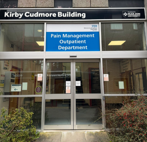 External double doors leading to building with signs overhead showing Pain Management Outpatient Department on NHS blue sign