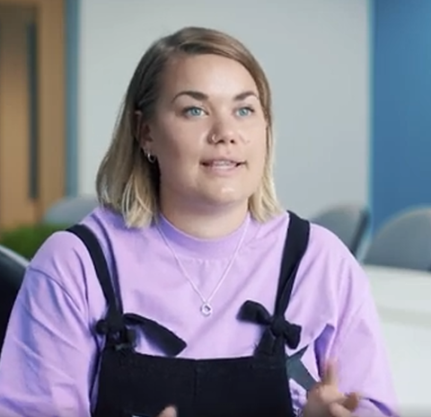 Young woman with blond bob, lilac t-shirt and dungarees talking to camera