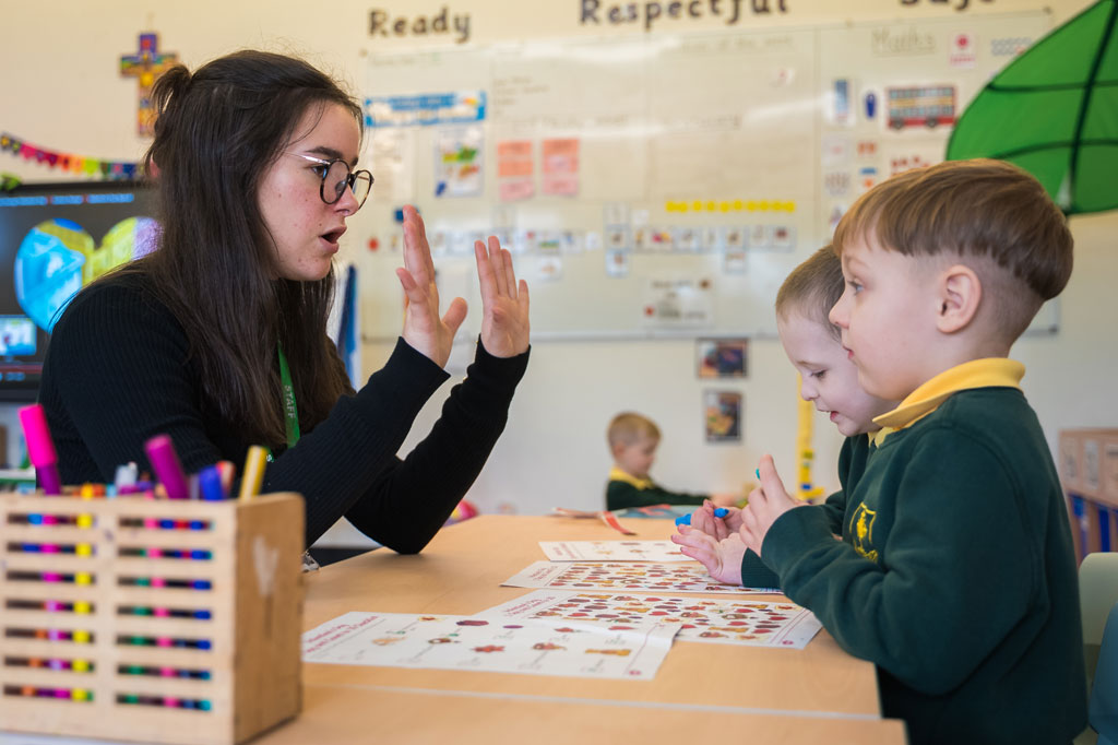 Woman teacher sat at classroom table with young boys in uniform sat opposite her