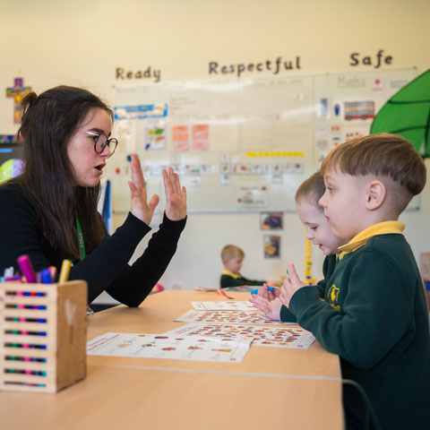 Woman teacher sat at classroom table with young boys in uniform sat opposite her